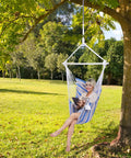 Person relaxing in a striped hammock chair with rope swing anti slip steel spreader bar, durable cotton ropes under a tree.