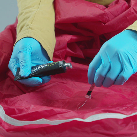 Person applying Seam Grip WP Waterproof Sealant And Adhesive For Jackets Tents Tarps Rain Gear And Outdoor Fabric Permanently Seals Seams And Repairs Rips On Nylon Canvas And Vinyl With A Flexible 1 Oz Tube That Covers Up To 12 Feet on a red fabric tear.