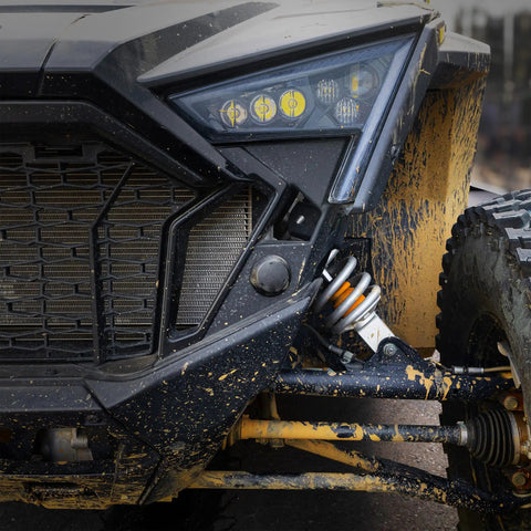 Close-up of muddy off-road vehicle front, showcasing headlight and suspension system, highlighting rugged terrain capabilities.