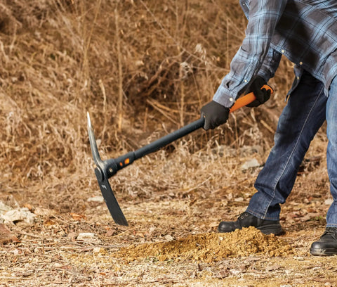 Person using IsoCore Dual Ended Pickaxe And Hoe With 36 Inch Shock Absorbing Handle for digging soil, showing orange and black tool in action.