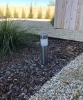 Single solar bollard light installed in garden bed showcasing Six Pack Solar Bollard Path Lights Outdoor Stainless Steel Warm White LED Landscape Lighting For Backyard Lawn Patio Waterproof Durable Silver Finish With Easy Ground Install.