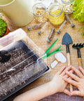 Child's hands with Six Pack Seed Starter Tray Kit With Humidity Domes, garden tools, seeds, and plants on table
