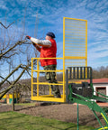 Forklift safety cage, yellow metal, mounted on a green aerial lift with a worker in a red vest trimming branches.