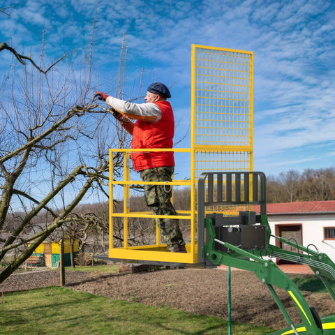 Forklift safety cage, yellow metal, mounted on a green aerial lift with a worker in a red vest trimming branches.