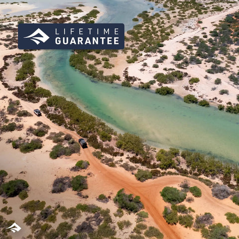 Aerial view of vehicles on sandy trail beside turquoise river, showcasing natural landscape for rugged adventures, lifetime guarantee badge.