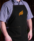 Person wearing black apron with flame logo, part of the Professional 22 Piece Griddle And BBQ Tool Kit Stainless Steel Dishwasher Safe For Quick Cleanup And Built To Last For Novice And Pro Grill Masters At Home And On The Grill.