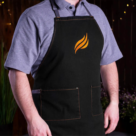 Person wearing black apron with flame logo, part of the Professional 22 Piece Griddle And BBQ Tool Kit Stainless Steel Dishwasher Safe For Quick Cleanup And Built To Last For Novice And Pro Grill Masters At Home And On The Grill.
