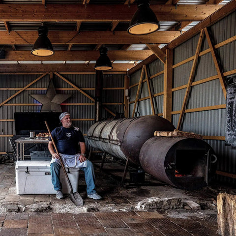Rugged Rotomolded Outdoor Cooler With Three Inch Insulation And Bear Proof Certification beside a man resting near a large smoker.