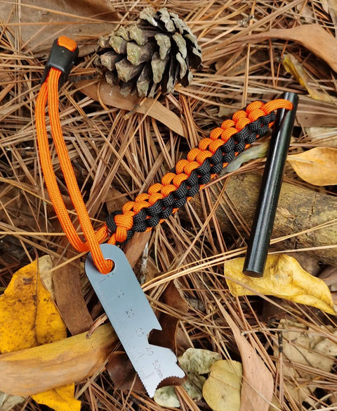 Drilled Ferro Rod Fire Starter Kit with 4 inch flint steel, paracord lanyard, striker, and whistle on a forest floor.