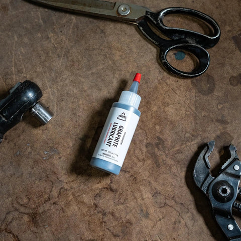 Powdered Graphite Lubricant 1.13 oz bottle on workbench with metal tools, for smooth sliding of locks, reels, and metal surfaces.