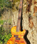 Four-string electric bass guitar with orange quilted maple top leaning against a tree outdoors.