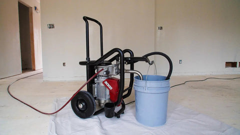 Professional Electric Airless Paint Sprayer Boasting PermaStroke Tech And 3300 Psi Thrust next to a blue paint bucket on a cloth floor.