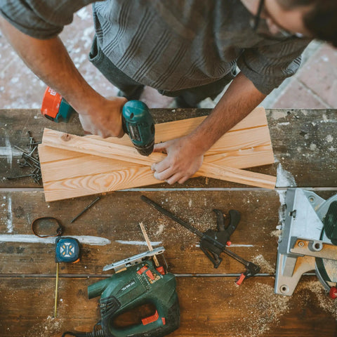 Person using electric drill on light wood plank in workshop surrounded by tools like saws, clamps, and measuring tape.