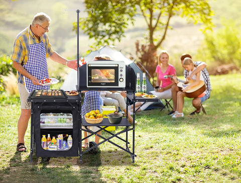 All In One Camping Kitchen Station With Light Stand And Windshield Portable Foldable Grill Table And Cook Station Picnic Table For Beach And Outdoors Includes Organizers And Carry Bag Black in use at campsite with people cooking and relaxing.
