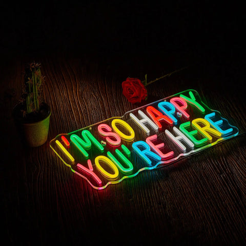 Colorful neon sign displaying 'I'm So Happy You're Here' on a dark wooden background, with a cactus and a red rose nearby.