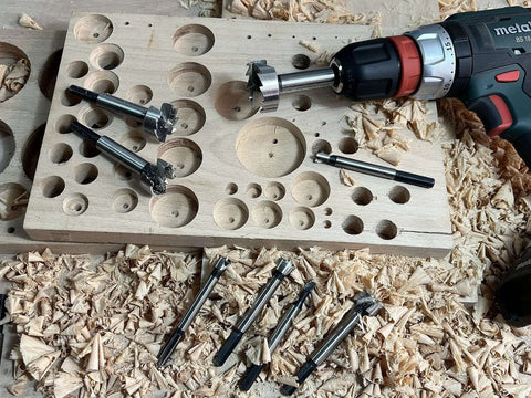 Overhead view of CORTOOL 16-piece Forstner bit set with hex shanks beside a Metabo drill on wood shavings.