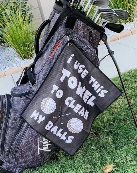 Golf bag with funny microfiber golf towel, 'Clean My Balls' joke, waffle-weave cleaning towel for clubs and balls, bag clip visible.