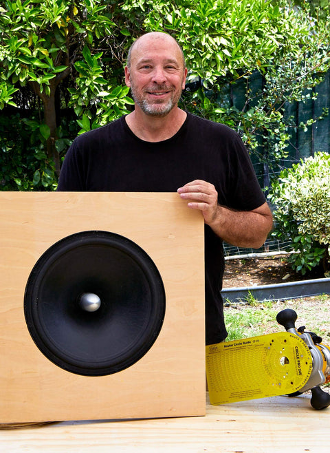 Man holding a wooden speaker cabinet with large black speaker outdoors, skateboard and greenery in the background.