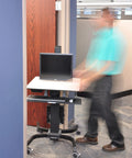 Smooth Roll Single Monitor Sit Stand Desk With Zero Effort Height Lift And Heavy Duty Caster Wheels For Vesa Monitors Up To Twenty Four Inches in an office setting with a person walking by.