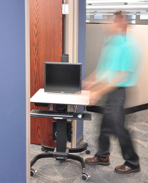 Smooth Roll Single Monitor Sit Stand Desk With Zero Effort Height Lift And Heavy Duty Caster Wheels For Vesa Monitors Up To Twenty Four Inches in an office setting with a person walking by.