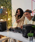 Two DJs using DM-50D Active 5-Inch Desktop Monitor Speakers, sleek black finish, for home studio and club DJ sets with crisp sound.