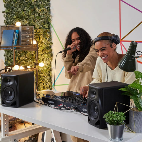 Two DJs using DM-50D Active 5-Inch Desktop Monitor Speakers, sleek black finish, for home studio and club DJ sets with crisp sound.