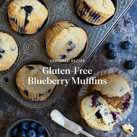 Baked gluten-free blueberry muffins with one sliced, buttered muffin on a rustic surface, showcasing wholesome texture and berries.