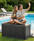 Two people sit on a black 90 Gallon Outdoor Deck Box With Wheels Lockable Waterproof Weatherproof Resin Storage Chest by a pool.