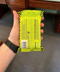 Hand holding a pack of On The Go Tech Wipes Pack Of 3, 25 Count Each, Travel Size Skin Safe Clean For Screens Phones Keyboards Monitors TVs And More, Safe For All Devices, Perfect For Home Office Or Travel, showing back view with yellow packaging.