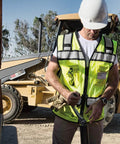 Rock Solid Hi Vis Surveyor Vest With Zipper Front Closure Ansi Class Two Type R Certified Lime Polyester Reflective Safety Vest Featuring Kickass Stripes And Hefty Tool Pockets For Xl worn by worker using tools on job site.