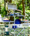 Portable Grill Table With Tons Of Prep Space For Camping, Tailgating, Picnics, And Boating in a forest setting with utensils and dishes