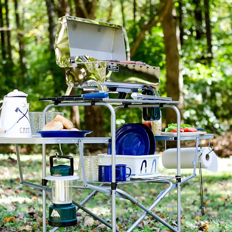 Portable Grill Table With Tons Of Prep Space For Camping, Tailgating, Picnics, And Boating in a forest setting with utensils and dishes