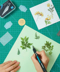 Person using Ergonomic Craft And Hobby Knife With 120 Replacement Number 11 Blades For Precision And Safety Ideal For Leather Scrapbooking Foam Clay Carving And DIY Arts And Crafts on green paper with leaf designs.