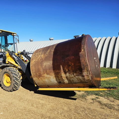 Forklift with yellow pallet forks lifting a large rusted steel drum outdoors