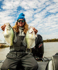 Angler on a boat holding two large crappie fish with a scenic river backdrop under a partly cloudy sky.