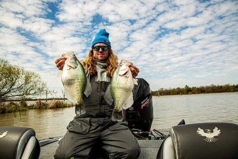 Angler on a boat holding two large crappie fish with a scenic river backdrop under a partly cloudy sky.