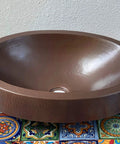 16 By 13 Inch Oval Skirted Vessel Copper Sink Hand Hammered Pure Copper Brown Finish For Modern Bath Semi Recessed Vessel One And A Half Inch Drain Opening Eighteen Gauge Handmade In Mexico CPS10 on colorful tile surface