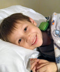 Boy in bed wearing green bedwetting alarm sensor clip under blanket, smiling on pillow