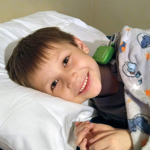 Boy in bed wearing green bedwetting alarm sensor clip under blanket, smiling on pillow