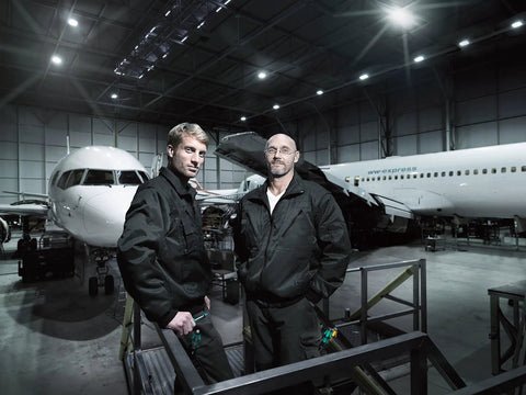 Two aircraft technicians standing in a hangar with two large commercial airplanes during maintenance.