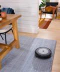 A Roomba vacuum on a rug in a living room with a wooden table, showcasing Three Pack High Efficiency Replacement Filters For Roomba E I J Series And Roomba Combo I5 J5 Traps Ninety Nine Percent Pet Dander Allergens And Keeps Your Air Fresh.