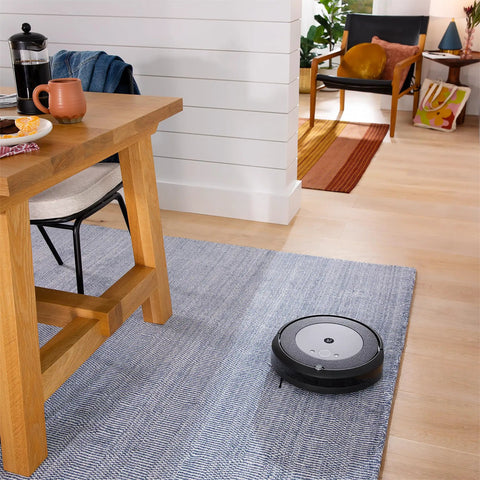 A Roomba vacuum on a rug in a living room with a wooden table, showcasing Three Pack High Efficiency Replacement Filters For Roomba E I J Series And Roomba Combo I5 J5 Traps Ninety Nine Percent Pet Dander Allergens And Keeps Your Air Fresh.