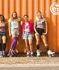 Four women in casual wear and roller skates stand by an orange container, smiling outdoors.