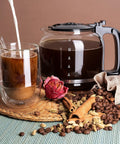 A filled borosilicate glass carafe next to a coffee cup surrounded by coffee beans, cinnamon, and a dried rose.