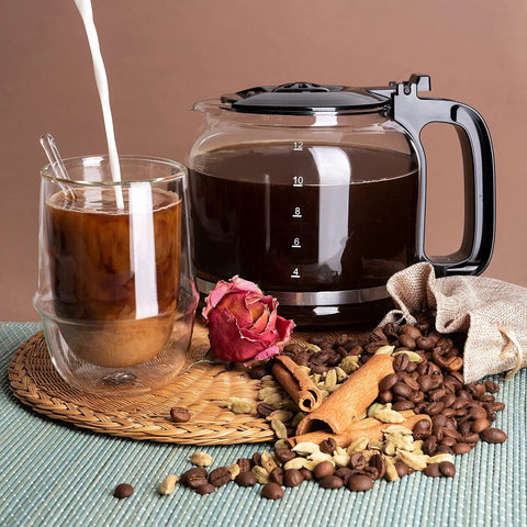 A filled borosilicate glass carafe next to a coffee cup surrounded by coffee beans, cinnamon, and a dried rose.