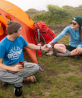 Two campers sitting by an orange tent with a red backpack, sharing a repair kit, outdoors on grass, wearing casual camping attire.
