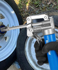 Close-up of KRETLAW mini grease gun with blue handle greasing a vehicle wheel hub via a grease fitting; metal head and spring.