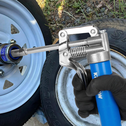Close-up of KRETLAW mini grease gun with blue handle greasing a vehicle wheel hub via a grease fitting; metal head and spring.