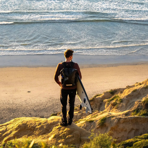 Person with surfboard wearing 25L Waterproof Surf Backpack For Adventure With Roll Top Closure at cliff overlooking beach