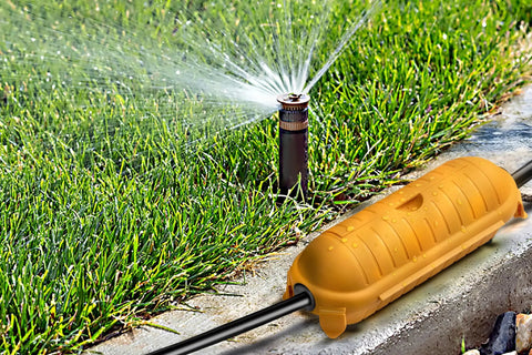Heavy Duty Outdoor IP44 Protective Case For Wired Connections Keeps Plugs And Sockets Dry Safe From Dirt And Damage Tough As Nails Weather Ready Rugged Housing For Extension Cords And Outdoor Equipment positioned by a garden sprinkler.